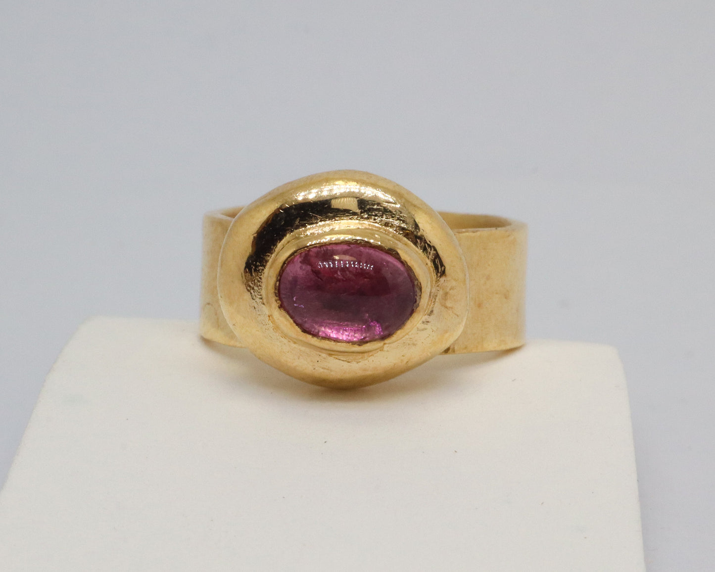 A gold ring with a textured oval bezel holding a round pink tourmaline gemstone, displayed on a white block against a light gray background.