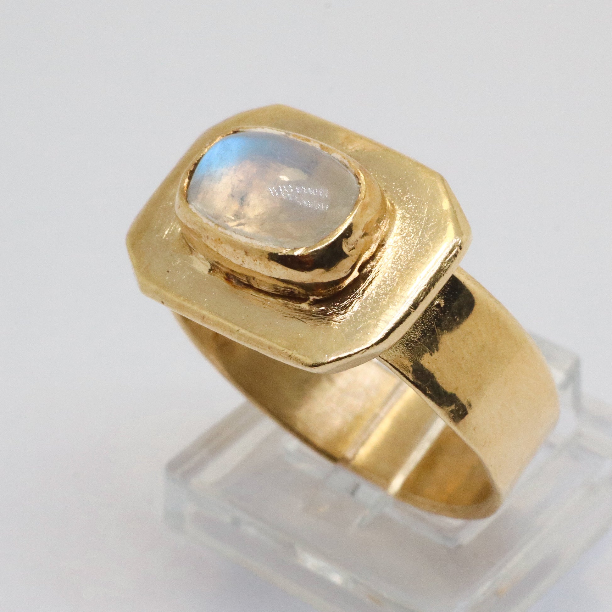 A gold signet ring with a bezel-set round iridescent moonstone, displayed on a clear acrylic stand against a white background.