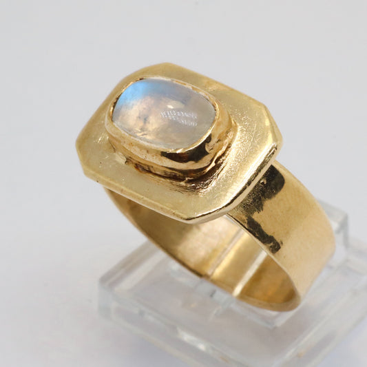 A gold signet ring with a bezel-set round iridescent moonstone, displayed on a clear acrylic stand against a white background.